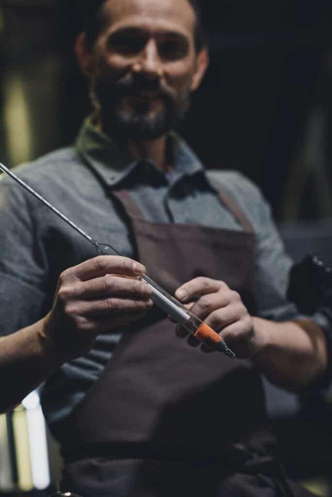 Smiling brewery worker in apron holding industrial thermometer while testing mead temperature