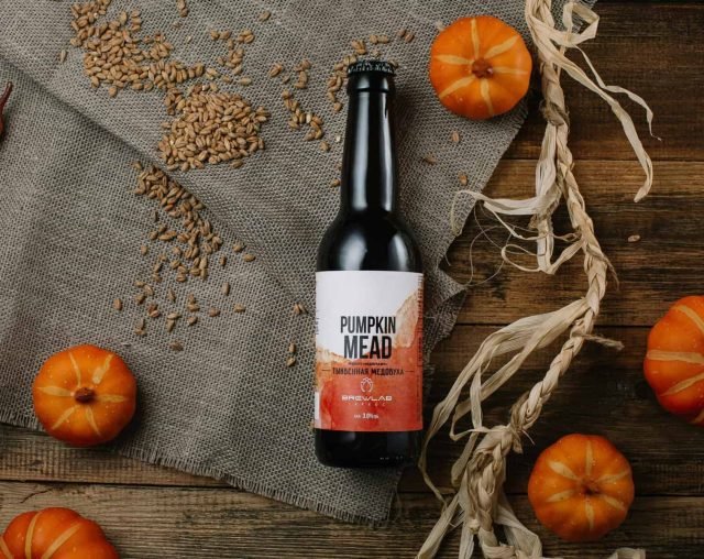 Bottle of pumpkin mead surrounded by small pumpkins and wheat grains on burlap.